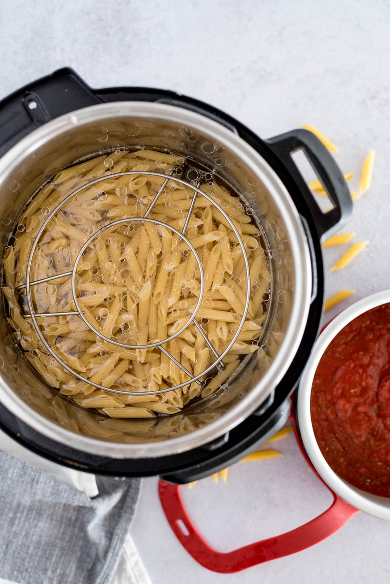 An overhead photo of the Instant Pot with the pasta and trivet inside and the tomato sauce in the silver cake pan, ready to sit on top of the trivet