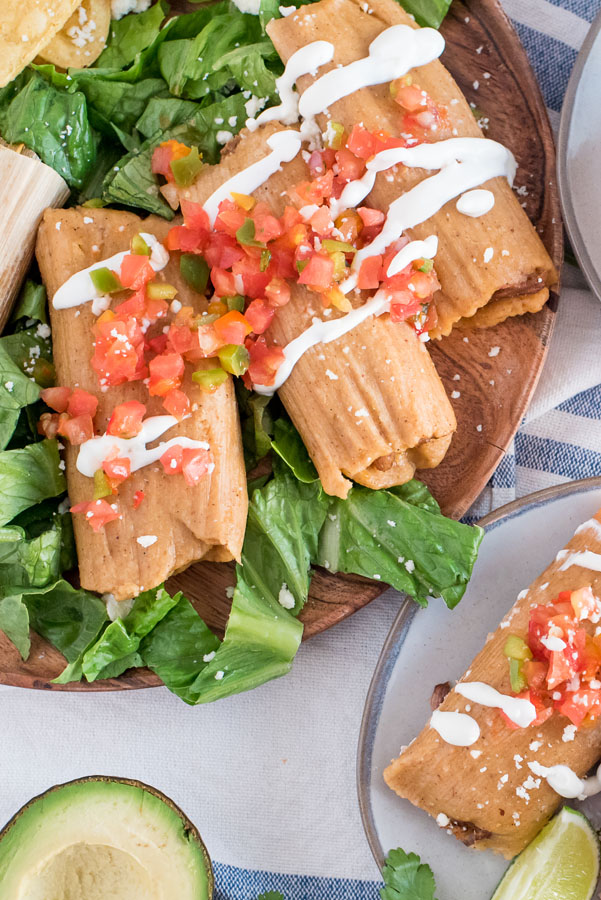 Overhead close up of several pork tamales, laying on a bed of lettuce, topped with tomatoes and sour cream, with an avocado in the bottom left corner