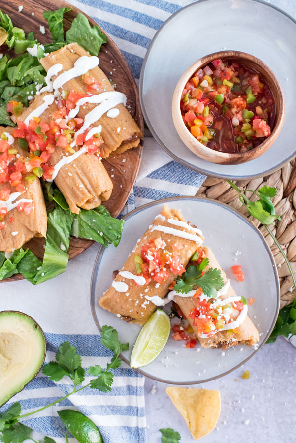 Overhead of a large serving platter and a smaller plate filled with homemade pork tamales served with fresh salsa in a small bowl, sour cream, cilantro, lime slices and crunchy lettuce on a light grey background.