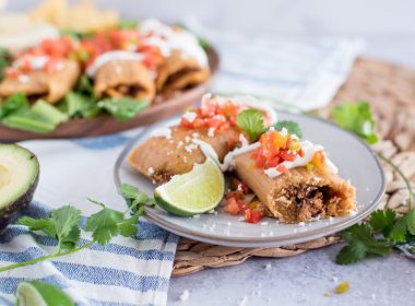 Homemade Pork Tamales wrapped in corn husks on a grey plate garnished with lime wedges, fresh salsa, cilantro, and cheese with half an avocado on the side.