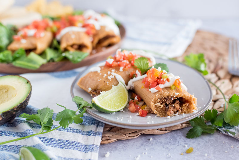 Homemade Pork Tamales wrapped in corn husks on a grey plate garnished with lime wedges, fresh salsa, cilantro, and cheese with half an avocado on the side.