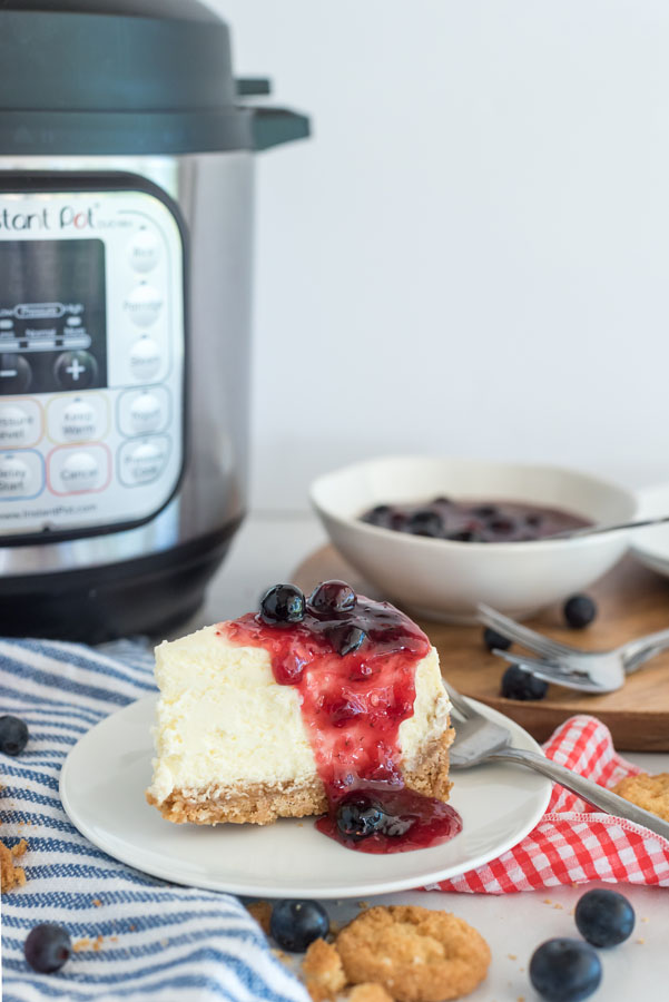 Side view of a slice of pressure cooker cheesecake topped with triple berry compote on a white plate, with an Instant Pot in the background.