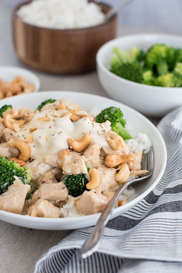 Close up of rosemary cashew chicken in a bowl with broccoli and rice, with bowls of broccoli, cashews, and rice in the background.