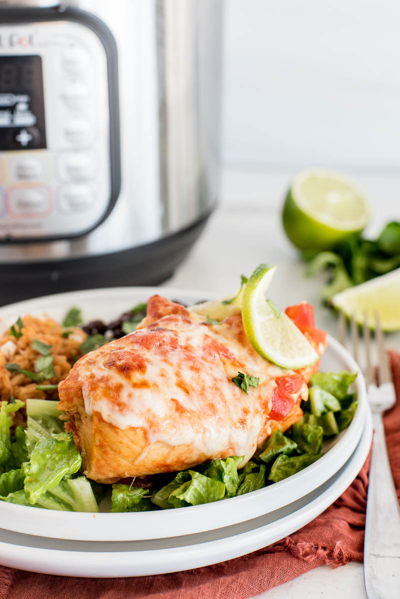 A close-up shot of Instant Pot Salsa Chicken, plated on a white plate, over a bed of shredded lettuce, with browned, melty cheese and a lime wedge on top, with an Instant Pot blurred in the background