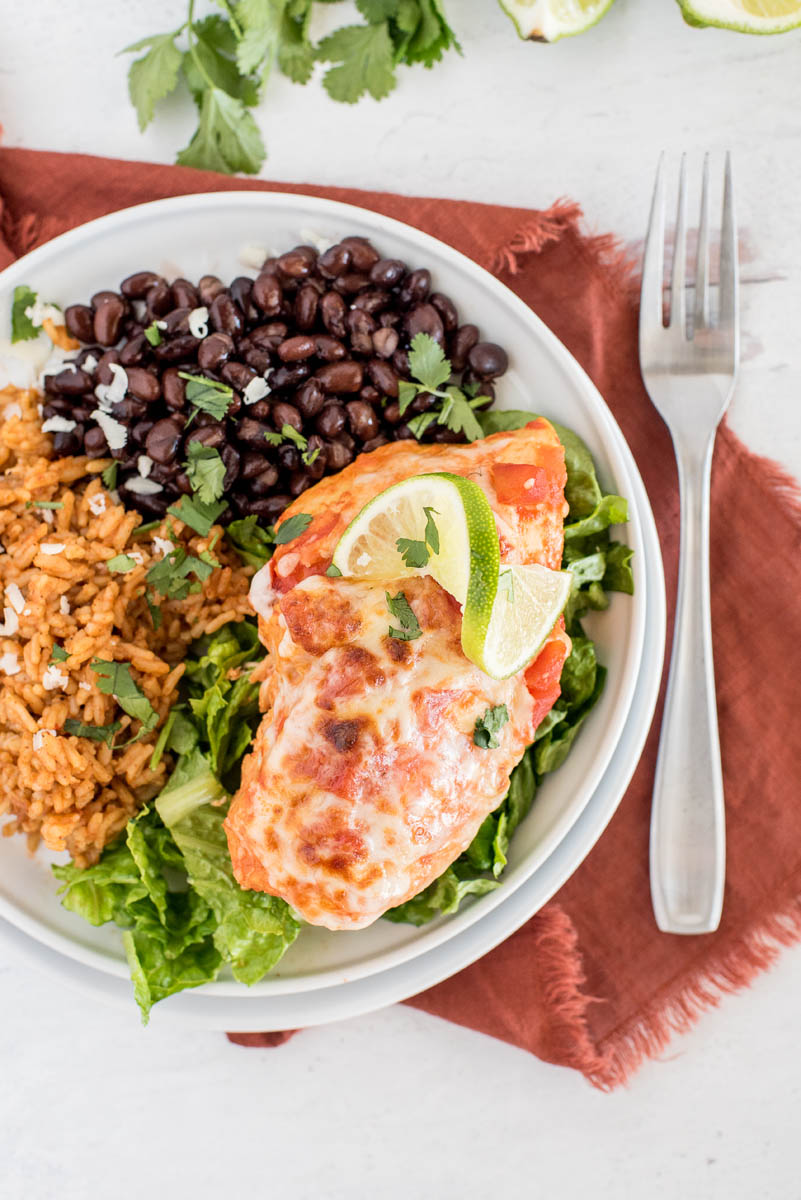 an overhead shot of pressure cooker Salsa Chicken, plated on a white plate garnished with a lime wedge and melty cheese, and served on top of a bed of shredded lettuce, with Mexican rice and black beans in the background