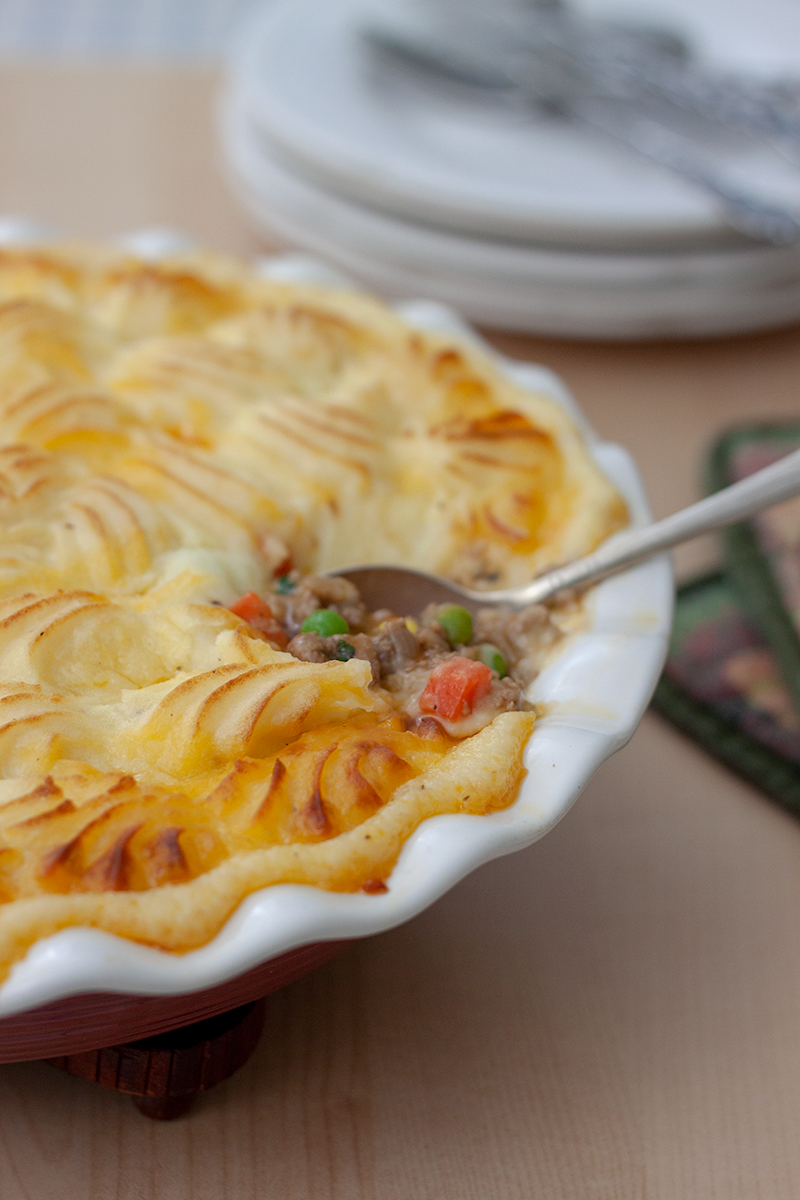 close shot of Instant Pot shepherds pie topped with mashed potatoes and browned in the oven, with a silver spoon showin gthe ground beef, carrot, and corn filling.