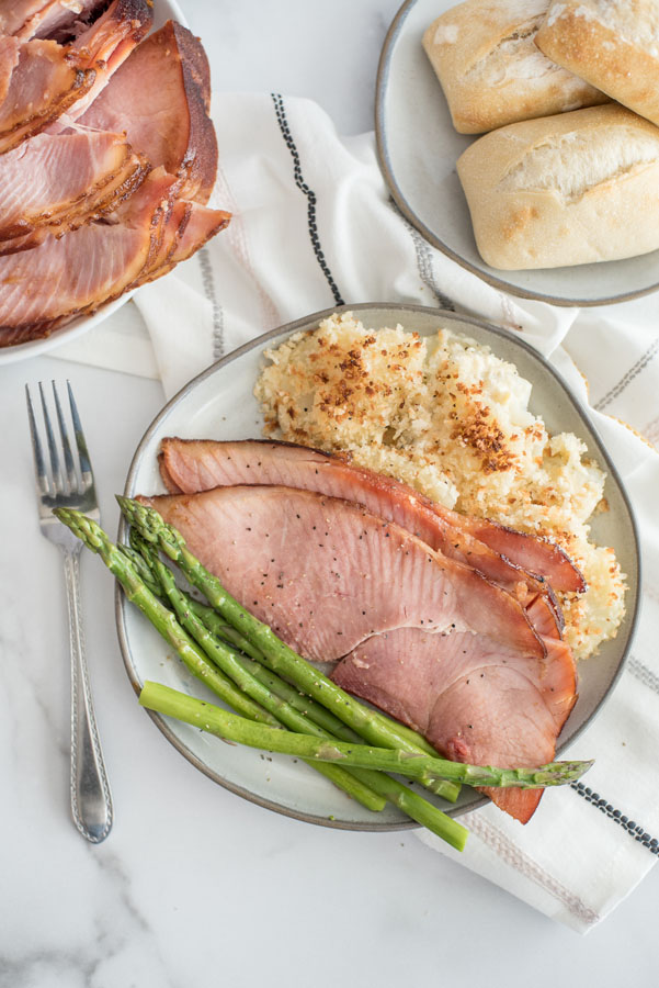 Overhead of a white plate filled with spiral ham slices, crisp asparagus, and crispy potato gratin