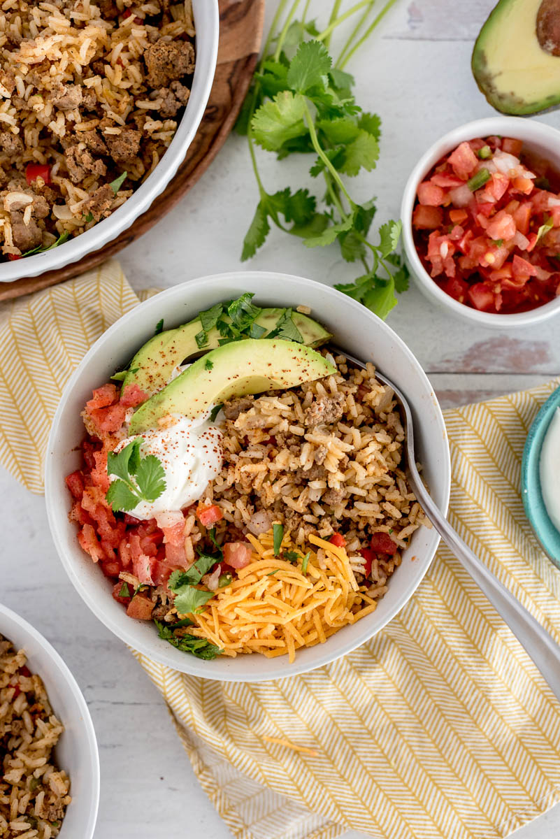 Overhead picture of Instant Pot taco rice topped with sliced avocado, cheese, cilantro, tomatoes, and sour cream. Placed in a white bowl with tomatoes, fresh cilantro, and a serving bowl with taco rice in the background.