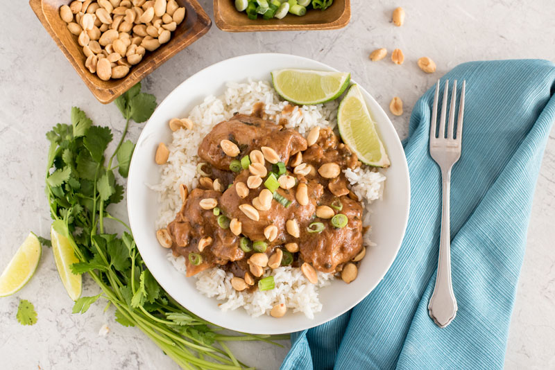 an overhead shot of Instant pot peanut thai chicken with cilantro lime sauce and white rice, with a blue napkin and a fork on it, a sprg on cilantro on the other side, and small dishes of peanuts and green onions at the top