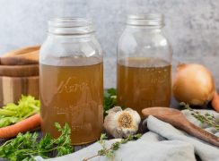 Side view of mason jars filled with Instant Pot turkey stock surrounded by fresh garlic, onion, thyme, parsley, and carrots.