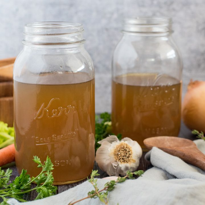 Side view of mason jars filled with Instant Pot turkey stock surrounded by fresh garlic, onion, thyme, parsley, and carrots.
