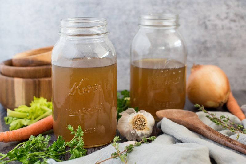 Side view of mason jars filled with Instant Pot turkey stock surrounded by fresh garlic, onion, thyme, parsley, and carrots.