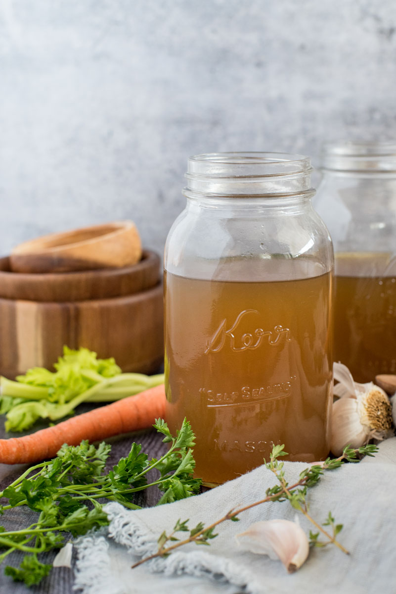 Side view of pressure cooker turkey stock on a mason jar surrounded by fresh garlic, thyme, parsley, carrots, and cellery.