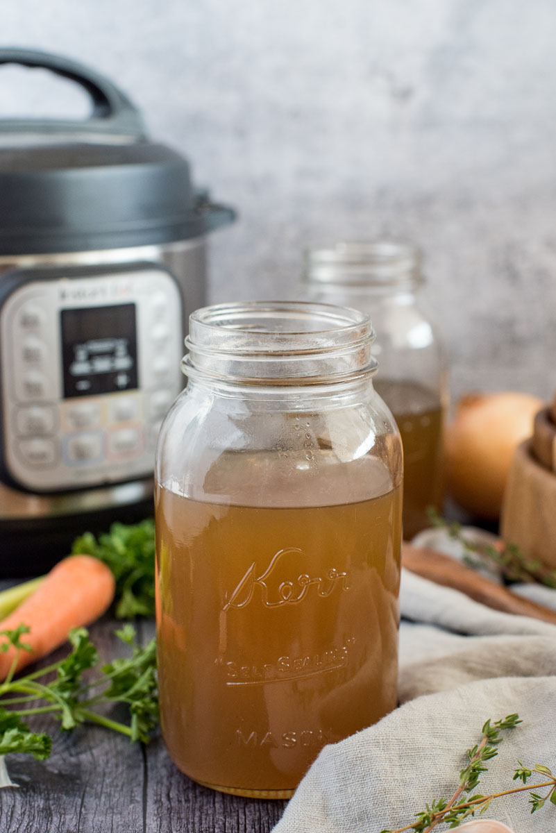 Insta Pot turkey stock in a mason jar in front of an Instant Pot, surrounded by fresh thyme, parsley, and carrots.