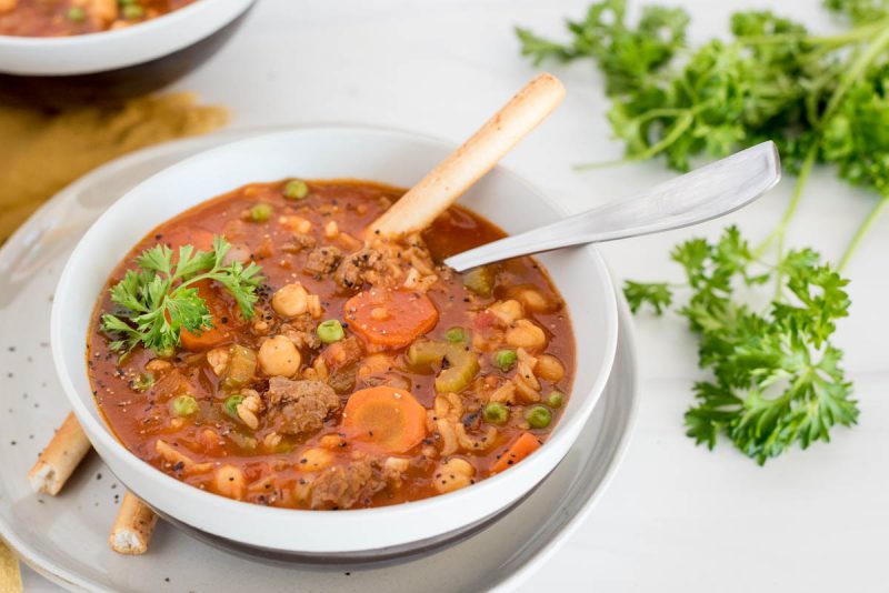 A close up picture of Instant Pot vegetable beef and rice soup garnished with fresh parsley and served in a white bowl with fresh parsley in the background.