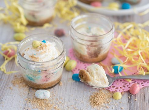 Side view of two mini pressure cooker Robin Egg’s Cheesecake made in mini glass mason jars with Whoppers Robin Eggs on a white wooden table decorated for Easter.