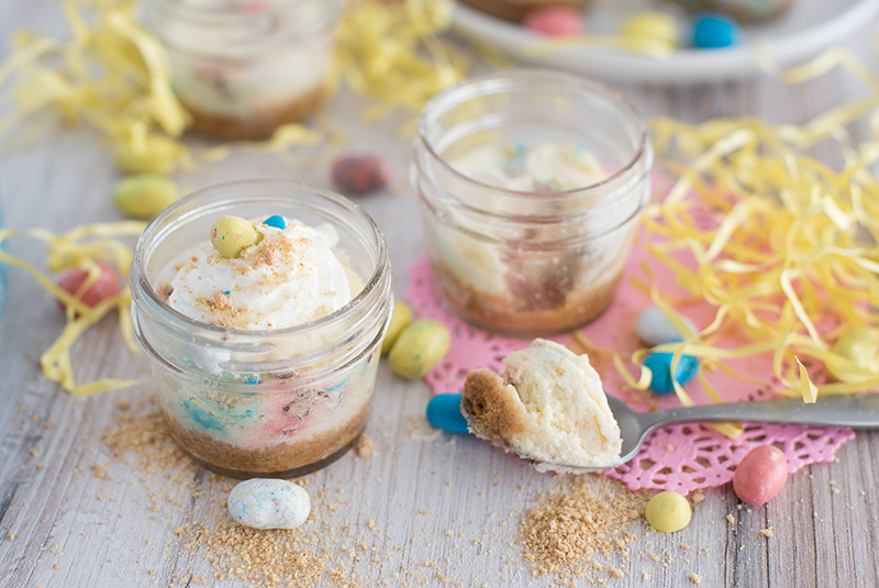 Side view of two mini pressure cooker Robin Egg’s Cheesecake made in mini glass mason jars with Whoppers Robin Eggs on a white wooden table decorated for Easter.