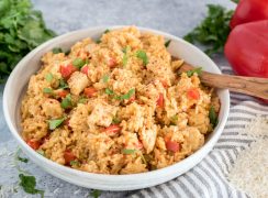 A 45 degree shot of a white bowl filled with Spanish style Instant Pot chicken and rice, garnished with red bell peppers and diced parsley, with a wooden spoon tucked in the back right of the bowl, with extra parsley and red bell peppers in the background