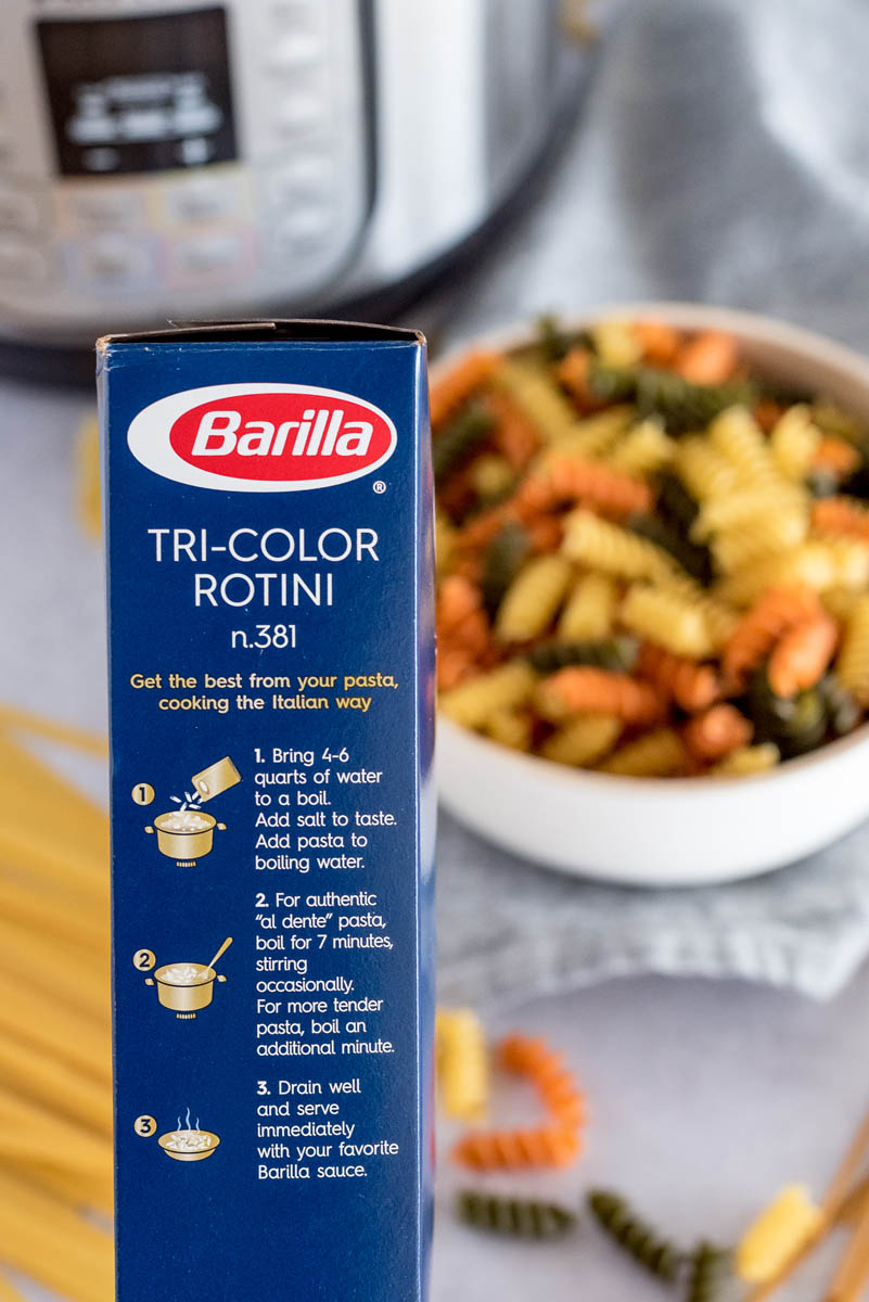 A close-up of the box of INstant Pot Tri-Color Rotini, with a bowlful of pasta and an Instant Pot in the background