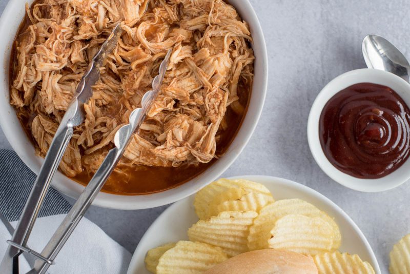 BBQ Chicken on a platter with metal tongs, with extra barbecue sauce in a small dish and potato chips on the bottom right of the shot