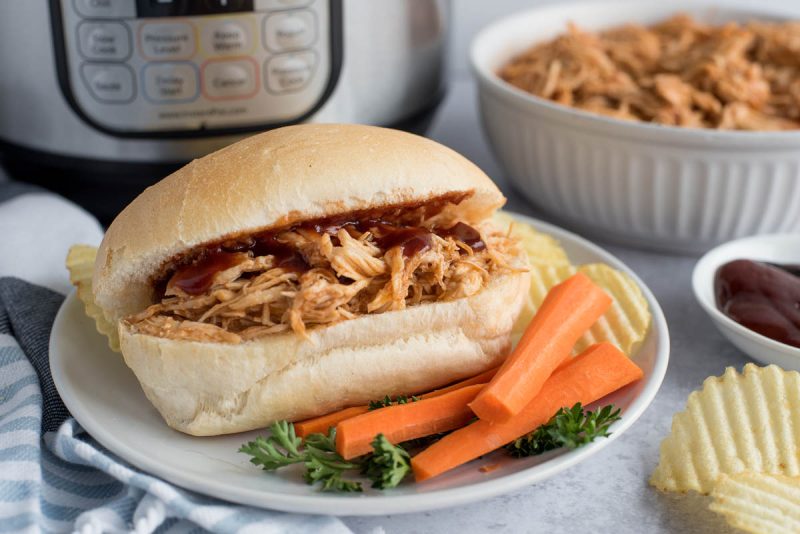 Pulled BBQ Chicken sandwich in front of an Instant Pot with carrots and potato chips plated in the foreground