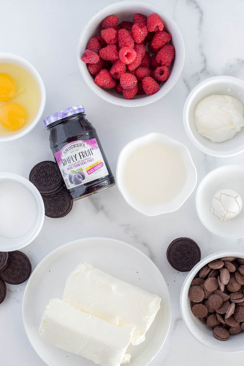 an overhead shot of the ingredients for Raspberry Cheesecake, including two eggs, fresh raspberries, sugar, flour, blocks of cream cheese, heavy cream, and melting chocolates