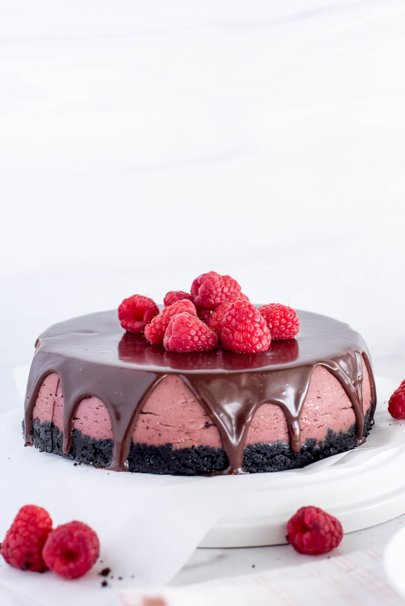 A beautiful centered shot of the finished raspberry cheesecake, covered in chocolate ganache with fresh raspberries on top.