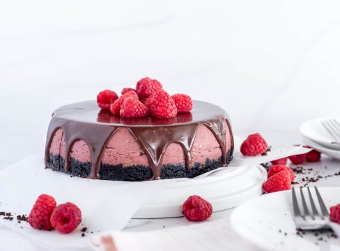 a side shot of a purple-red raspberry cheesecake with a chocolate cookie crust, topped with chocolate ganache drizzles and red raspberries. Sitting on a white plate with additional raspberries on the sides