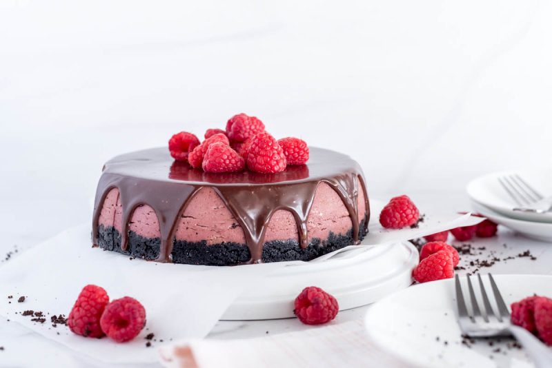 a side shot of a purple-red raspberry cheesecake with a chocolate cookie crust, topped with chocolate ganache drizzles and red raspberries. Sitting on a white plate with additional raspberries on the sides