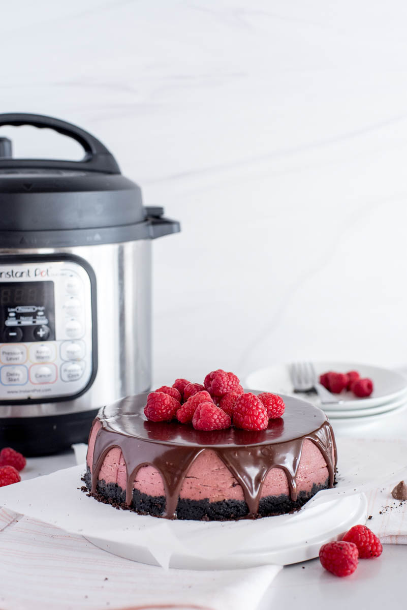 a chocolate raspberry cheesecake topped with fresh ganache and additional raspberries on a white platter in front of an Instant Pot and a stack of extra plates and forks with additional raspberries for garnish