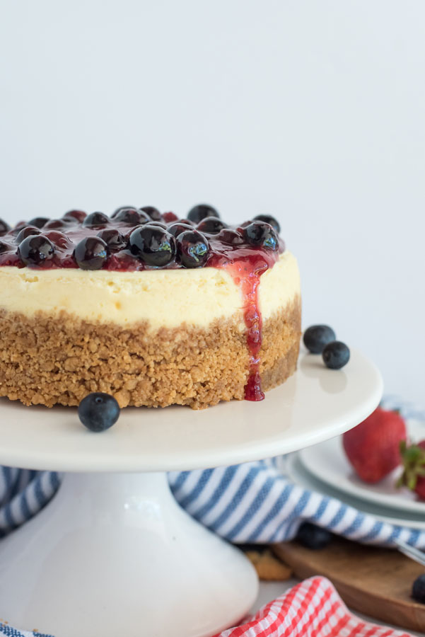 Side angle of Instant Pot cheesecake on a white cake stand, topped with triple berry compote and extra blueberries.