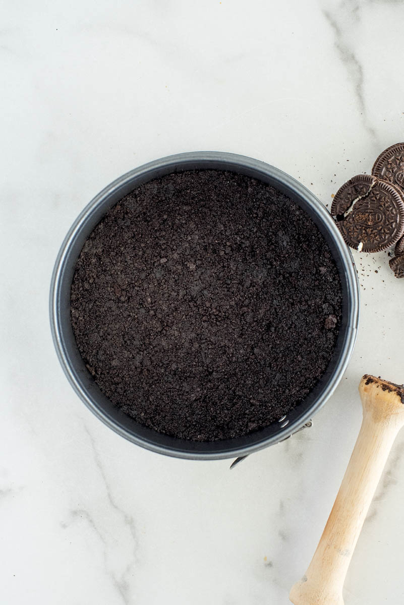 An overhead vertical shot of the chocolate cookie crumbs pressed flat in the springform pan, with the tamper and additional oreos visible on the outer edge.