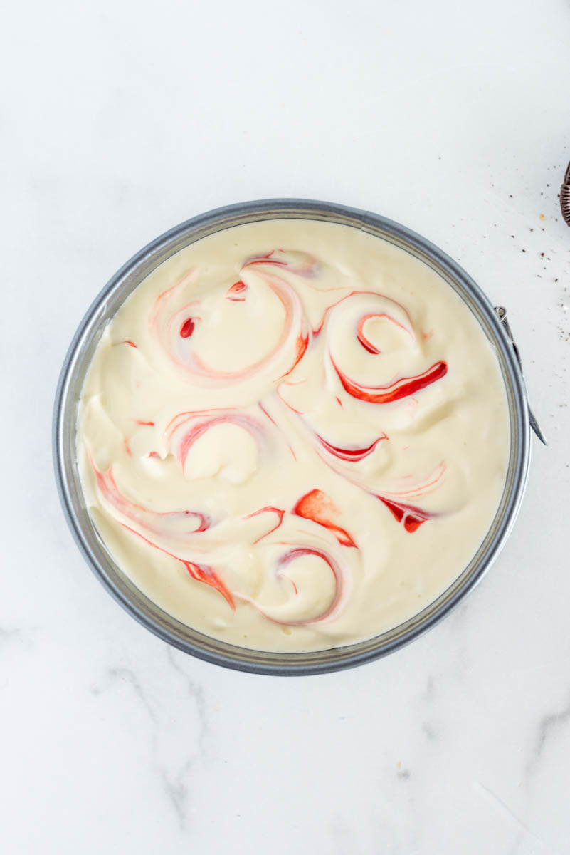 An overhead shot of a small springform pan showing the white layer poured on top and the red swirls twisted throughout.