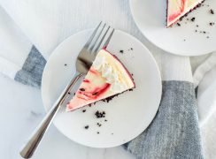 A slice of red velvet cheesecake on a small white plate with a fork, garnished with chocolate crumbs and finished with a whipped cream border