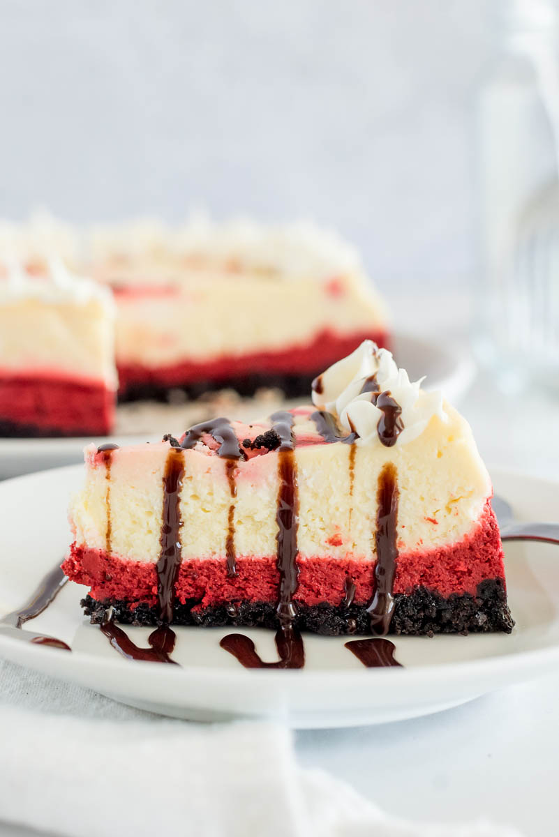 A side shot of Red Velvet Cheesecake, with the clear layers visible, drizzled in dark chocolate