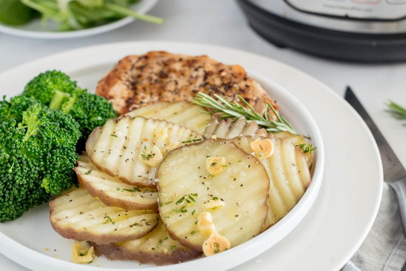 a close-up shot of the rosemary new potatoes garnished with salt, garlic, and rosemary, on a plate iwth broccoli and grilled chicken faded into the background