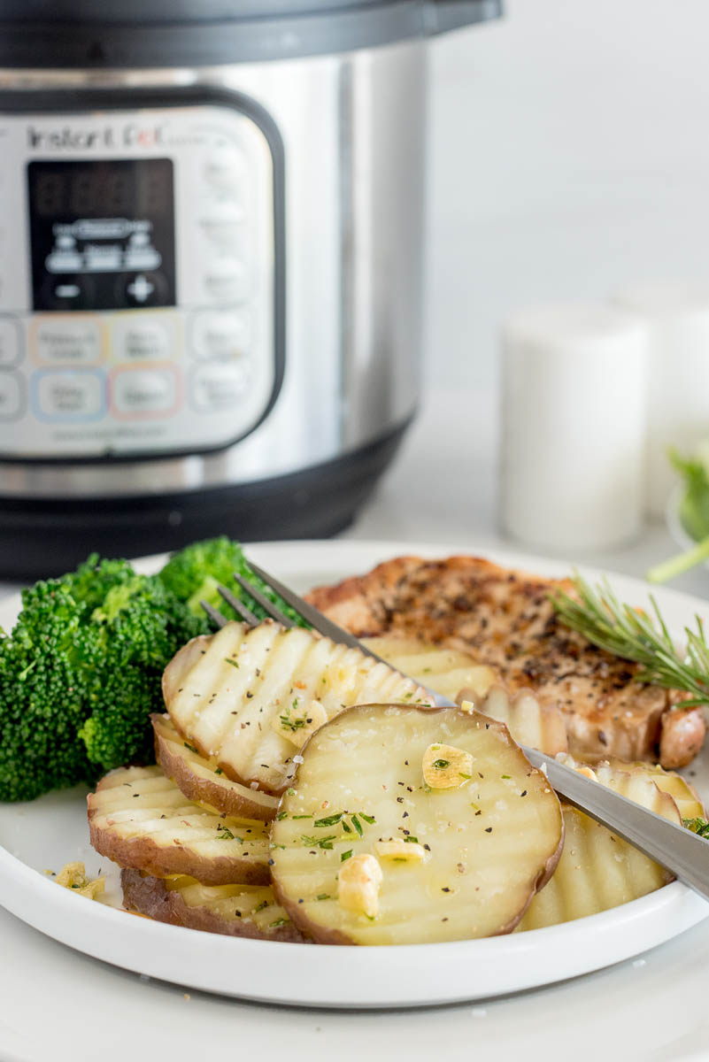 A 45-degreee shot of a white plate of rosemary new potatoes with a silver fork resting on top, with an out of focus Instant Pot in the background