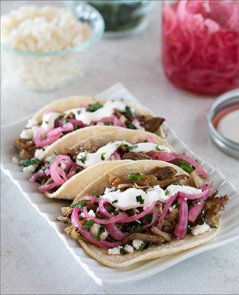 Pork Carnitas from the Electric Pressure Cooker Cookbook - featuring 45 degree shot of a white platter with three carnitas in a white corn tortilla with chopped cilantro, sour cream, and pink pickled onions garnished on topp