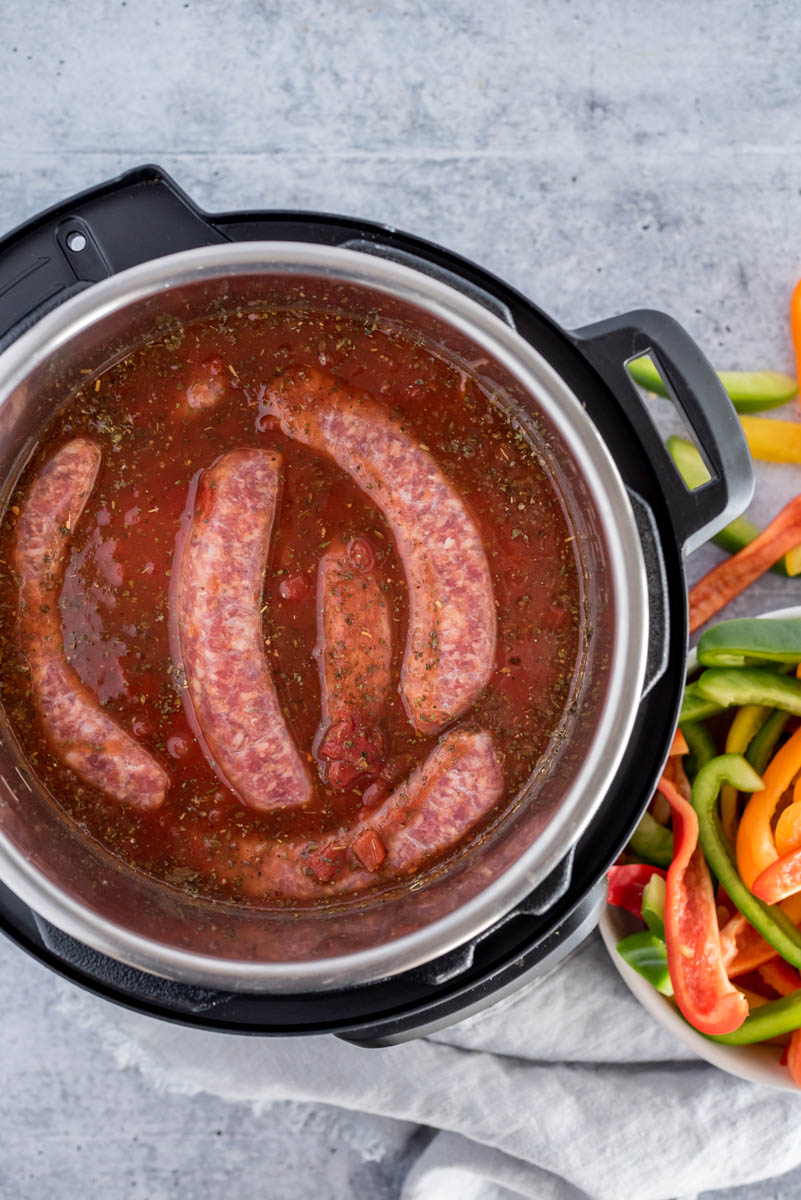 Uncooked sausages added to an Instant Pot with sliced peppers on the counter.