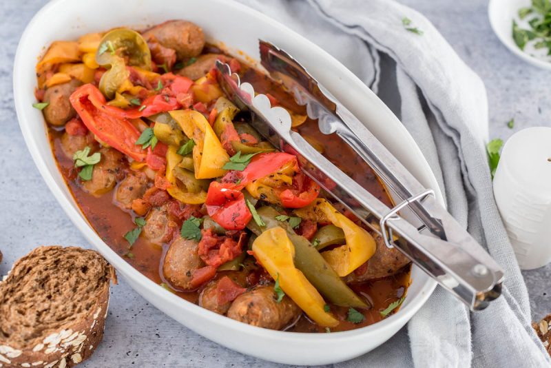 A white serving dish filled with Instant Pot sausage and peppers with tongs for serving, placed next to a gray napkin.