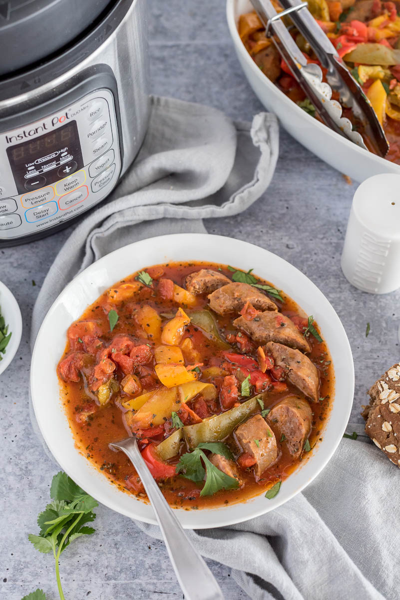 Overhead shot of a bowl of pressure cooker sausage and peppers in a white bowl, in futon of an Instant Pot and next to a white serving bowl.