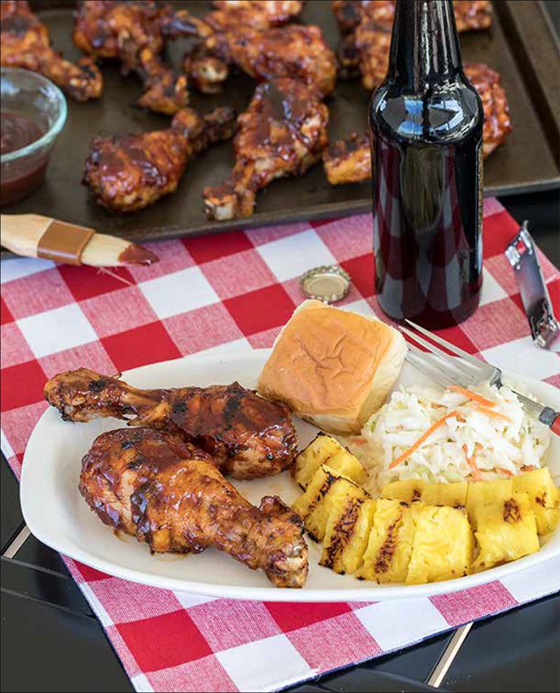 Quick Chicken Legs from the Electric Pressure Cooker Cookbook - featuring 45 degree shot of a white plate with two BBQ chicken legs, grilled pineapple, coleslaw, a roll, with a bottle of rootbeer and a dark sheet pan full of additional chicken legs in the background