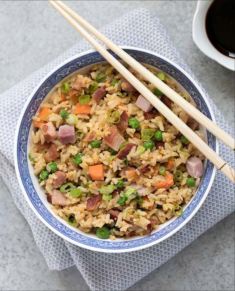 Ham Fried Rice from the Electric Pressure Cooker Cookbook - featuring overhead shot of yellow fried rice with bacon, carrots, ham, peas, fried egg, and green onions, with chopsticks balanced on top of the bowl and a small white bowl of soy sauce in the background