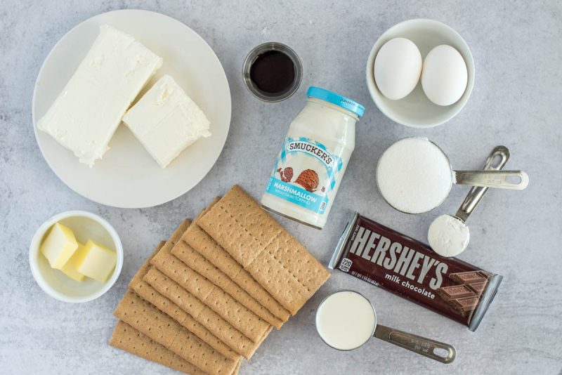 Ingredients for an Instant Pot s'mores cheesecake, including graham crackers, chocolate, sugar, heavy cream, eggs, marshmallow topping, cream cheese, butter, vanilla, and sour cream.