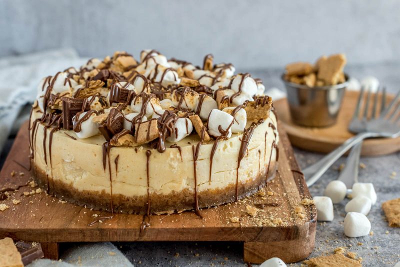 An Instant Pot smore cheesecake topped with marshmallows, graham crackers, and chocolate, placed on a wooden cutting board.