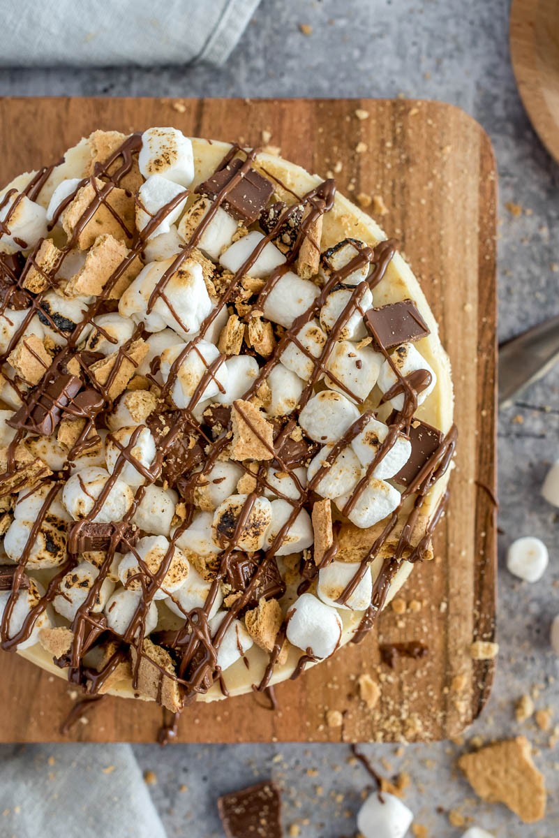 Overhead picture of an Instant Pot s'more cheesecake topped with marshmallows, graham crackers, and chocolate, placed on a wooden cutting board.