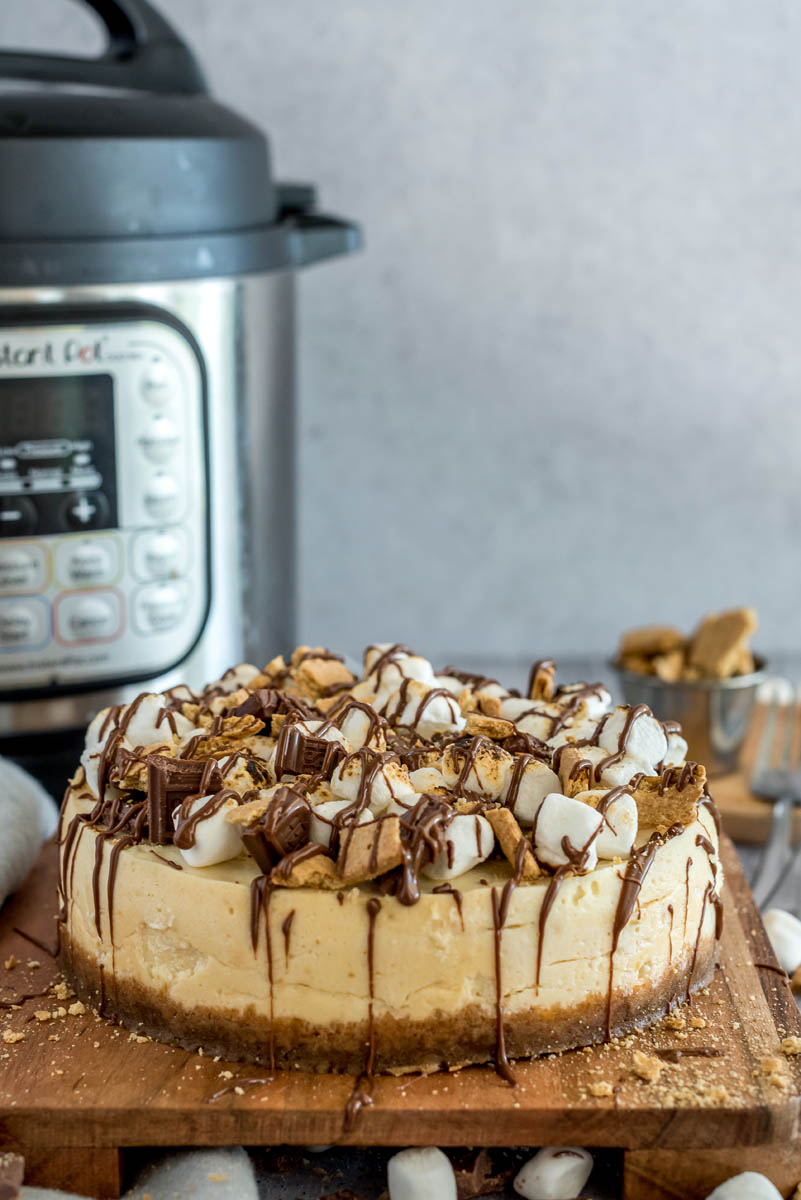 A s'mores cheesecake topped with marshmallows, graham crackers and chocolate, placed on a wooden cutting board and placed in front of an Instant Pot.