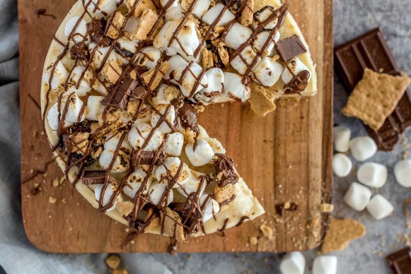 Overhead picture of an Instant Pot s'more cheesecake topped with marshmallows, graham crackers, and chocolate, placed on a wooden cutting board.