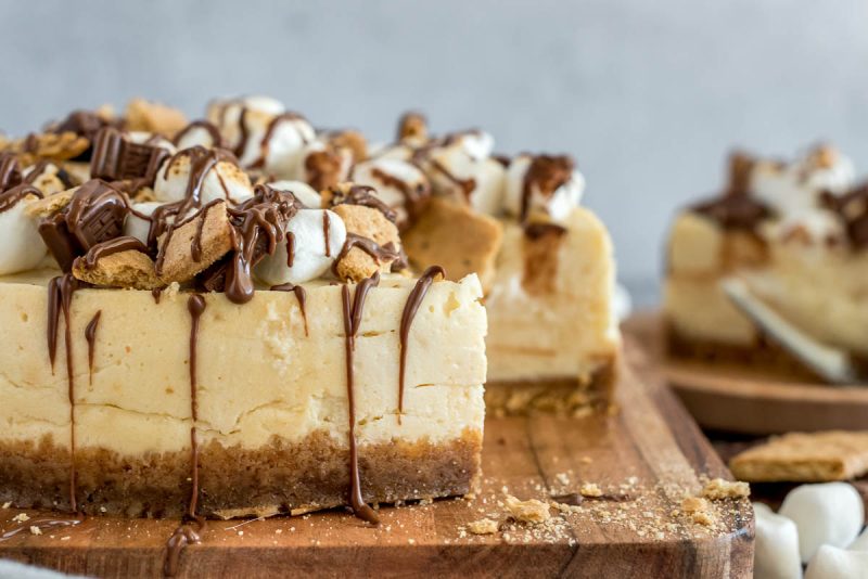 An Instant Pot s'mores cheesecake topped with marshmallows, graham crackers, and chocolate, placed on a wooden cutting board.