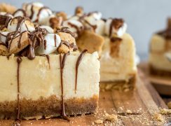 A very close-up shot of s'mores cheesecake topped with marshmallows, graham crackers, and melted Hershey's chocolate, placed on a wooden cutting board.